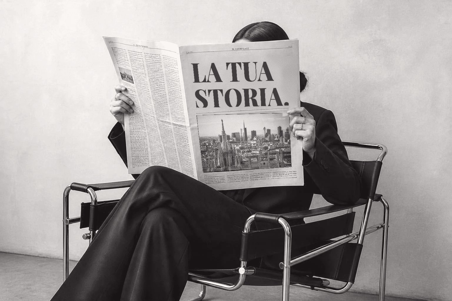 Donna in tailleur nero seduta in poltrona che legge un giornale con il titolo La tua storia, in bianco e nero